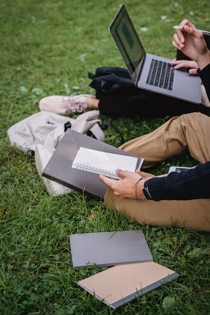 Two students on a lawn studying with a laptop and notebooks, focusing on coursework outdoors.
