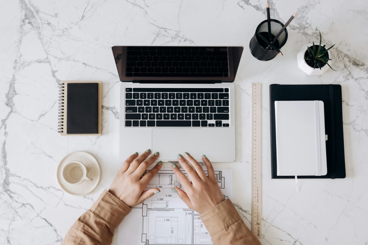 Top view of a stylish home office desk with a laptop, planner, and coffee cup, showing hands on a blueprint.